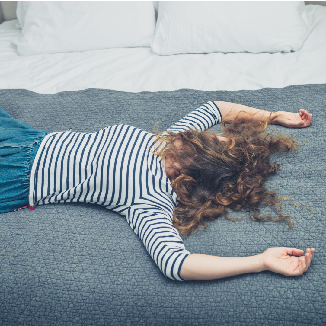 A woman with curly brown hair lies face-down on a bed, arms outstretched, wearing a striped shirt and denim skirt.